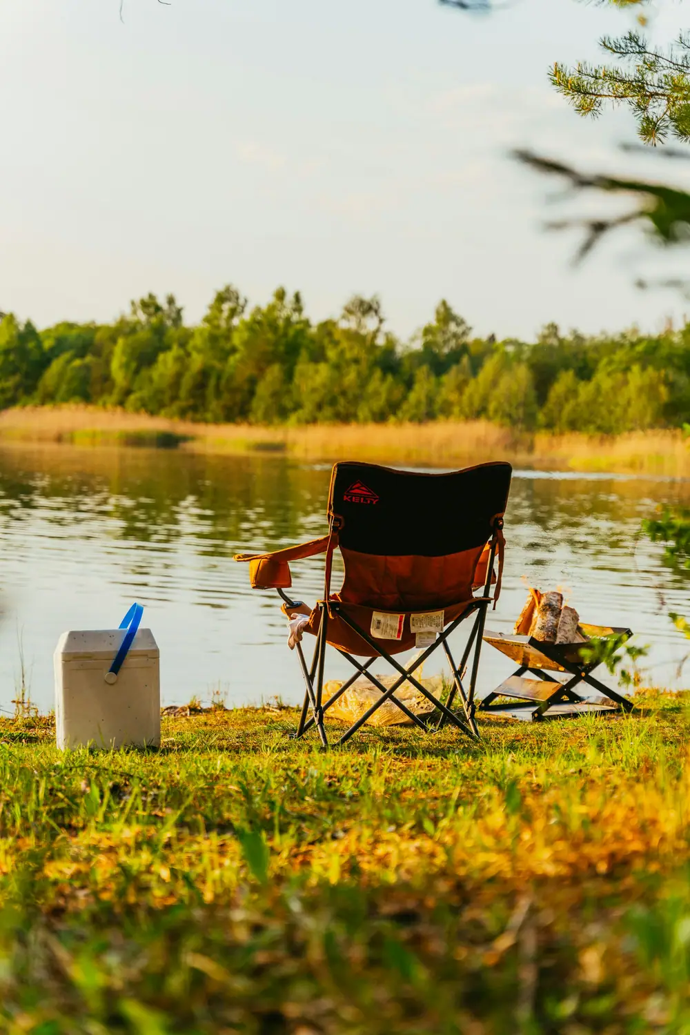A folding camp chair and small table set up on the grassy shore of a calm lake at golden hour