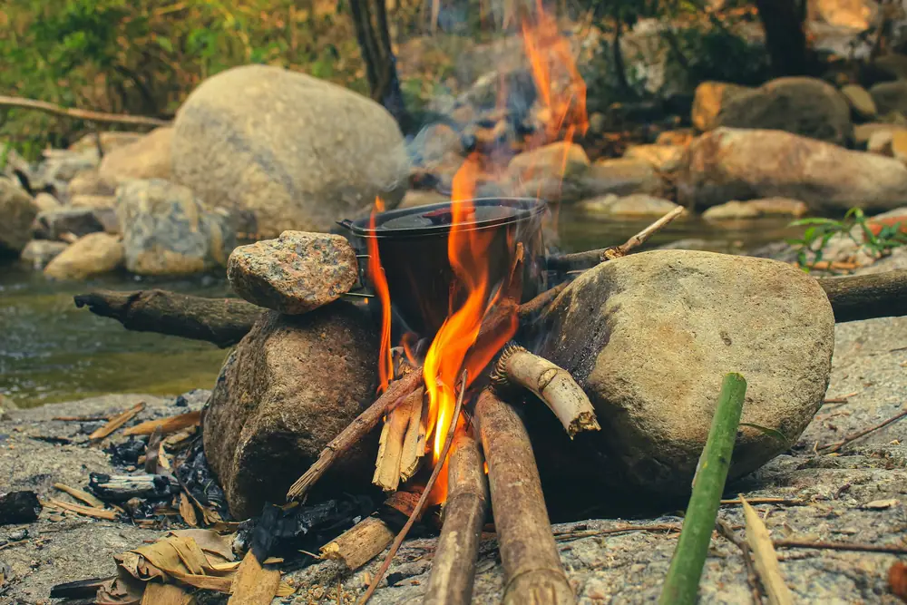 A blackened camp pot heating on a small wood fire between river stones