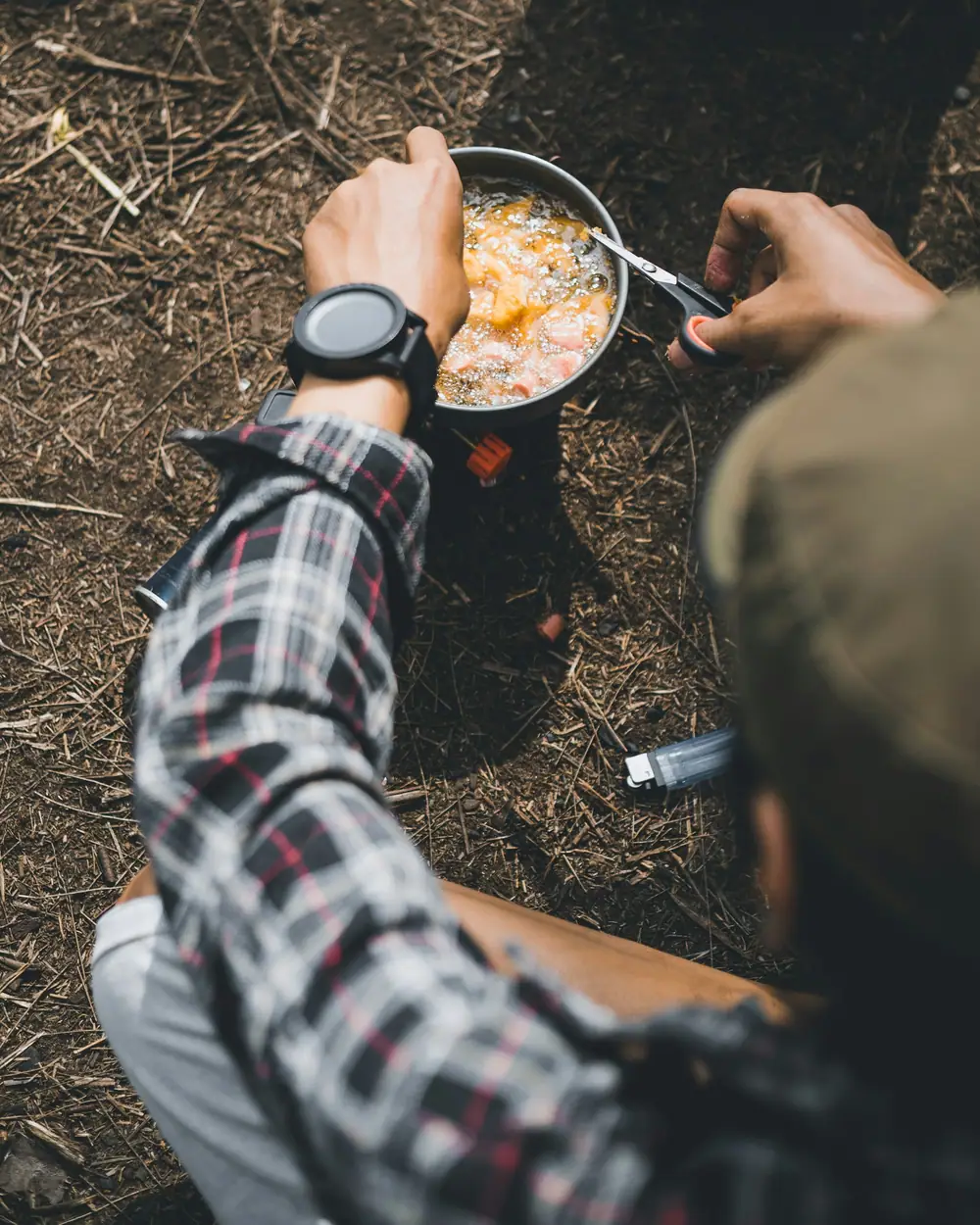 A camper in a flannel shirt stirring a pot on a small stove set on forest ground