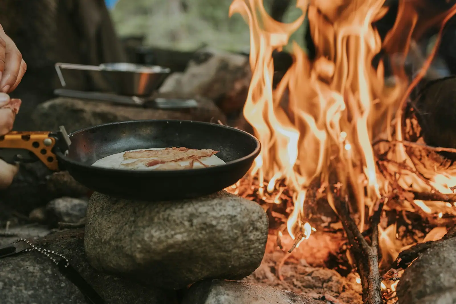 A skillet of food cooking on a flat stone beside an open campfire