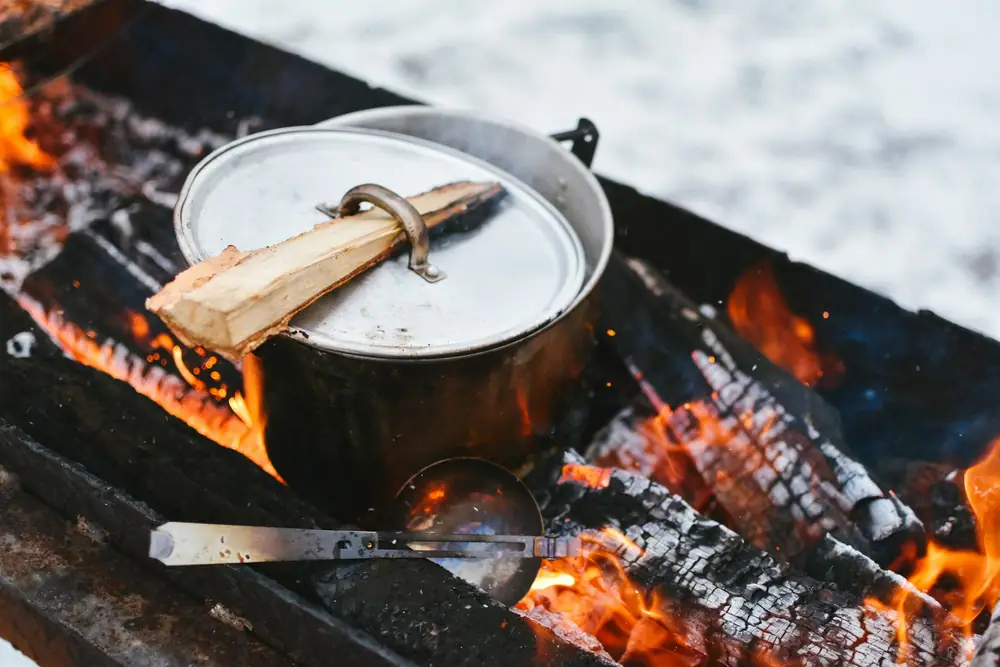 Covered steel pot heating water over a wood fire with a piece of firewood resting on the lid