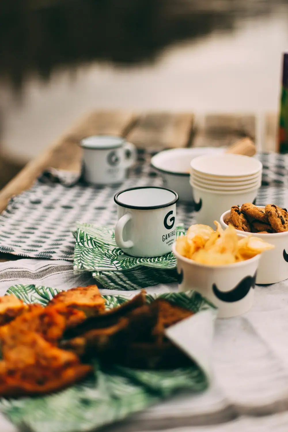 Enamel camp mugs, snack bowls and cups arranged on a gingham tablecloth at a campsite