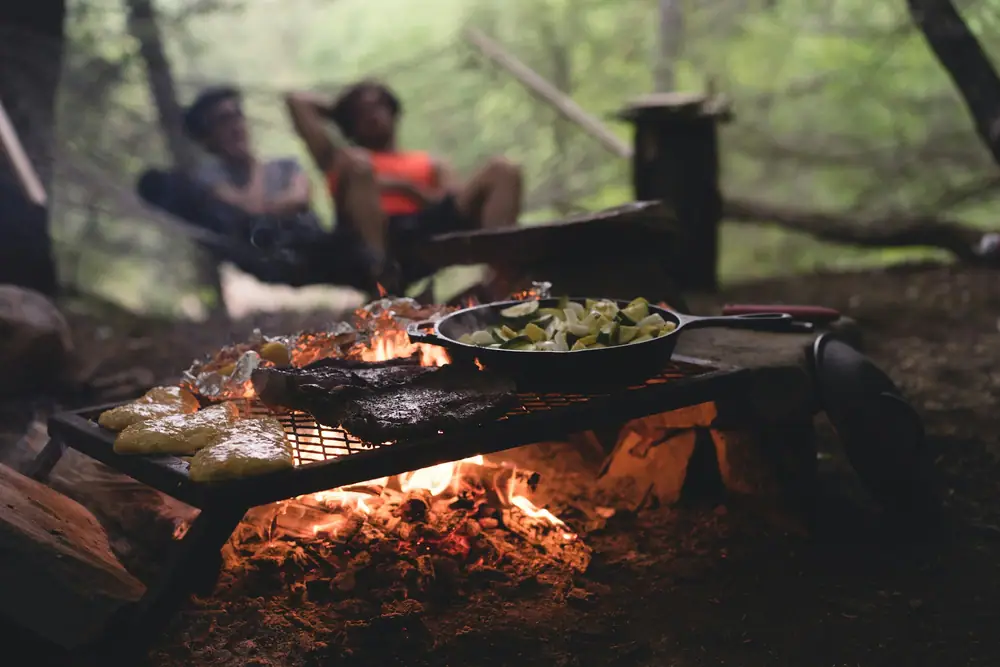 Food cooking on a grill grate and in a cast iron pan over glowing campfire coals in the evening