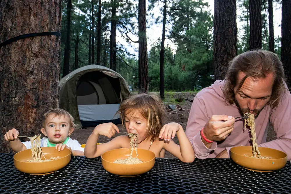 A father and two young children eating noodles from bowls at a picnic table in a forest campsite